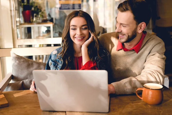 Hombre alegre mirando chica atractiva sonriendo cerca de la computadora portátil en la cafetería - foto de stock