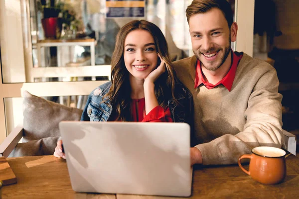 Hombre alegre y atractiva mujer joven mirando la cámara cerca de la computadora portátil en la cafetería - foto de stock