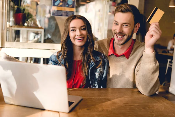 Весёлый мужчина держит кредитку рядом с счастливой молодой женщиной в кафе — стоковое фото
