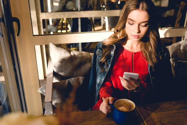 Vista aérea de atraente jovem segurando xícara de café e usando smartphone no café — Fotografia de Stock