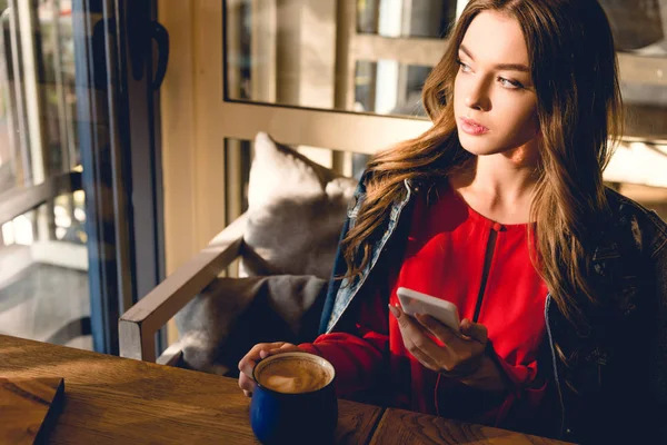 Atractiva mujer joven sosteniendo taza de café y teléfono inteligente en la cafetería - foto de stock