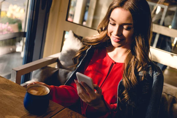 Щаслива молода жінка тримає чашку кави і дивиться на смартфон в кафе — стокове фото