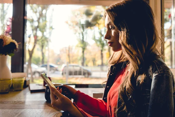 Привлекательная женщина держит чашку кофе и использует смартфон в кафе — стоковое фото
