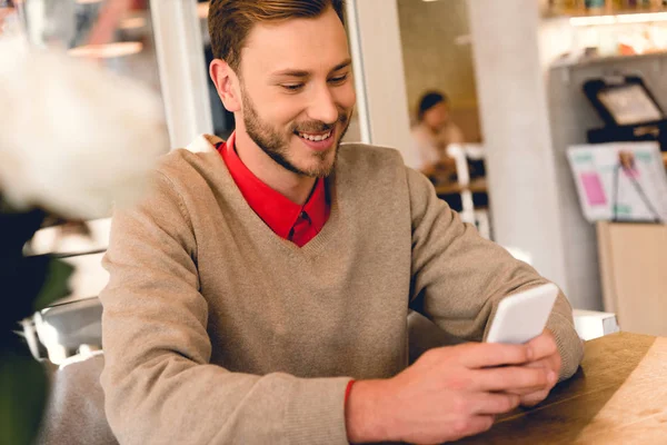 Красивый бородатый мужчина смотрит на смартфон и улыбается в кафе — стоковое фото