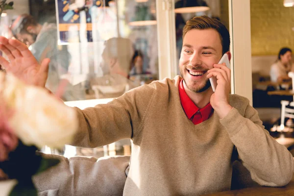 Hombre barbudo alegre agitando la mano y sonriendo mientras habla en el teléfono inteligente en la cafetería - foto de stock