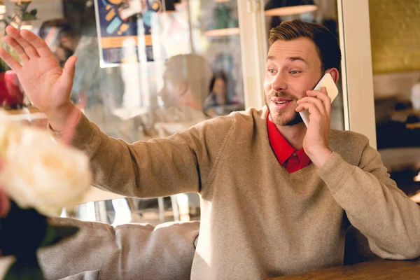 Hombre barbudo alegre saludando la mano mientras habla en el teléfono inteligente en la cafetería - foto de stock