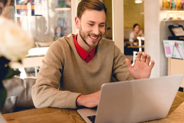 Happy blogger waving hand while having video chat on laptop in cafe — Stock Photo