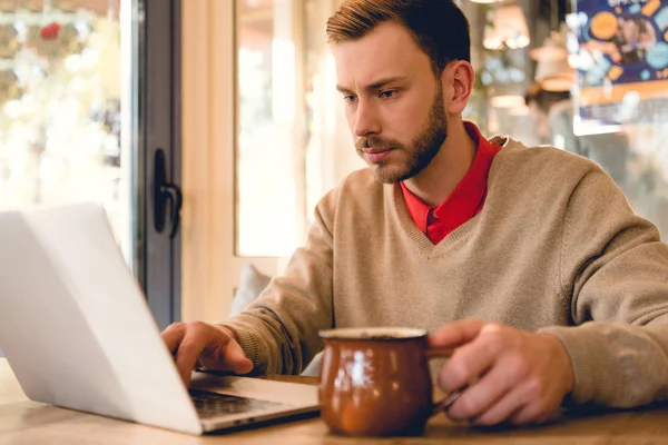 Bloguero concentrado mirando el portátil mientras sostiene la taza de café - foto de stock