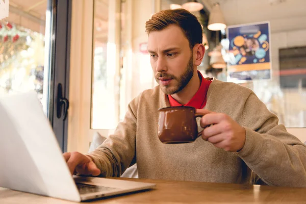 Guapo bloguero mirando portátil mientras sostiene la taza de café en la cafetería - foto de stock