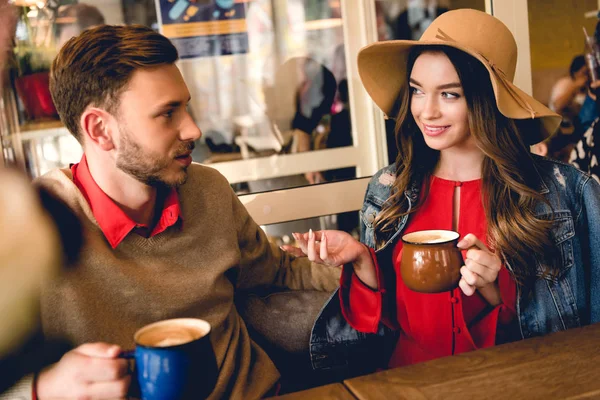 Schöne junge Frau mit Hut schaut Mann an, während sie eine Tasse Kaffee in der Hand hält — Stockfoto