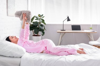 side view of woman in pink pajamas with teddy bear lying on bed at home clipart