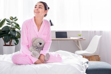 cheerful woman in pink pajamas with teddy bear sitting on bed at home clipart