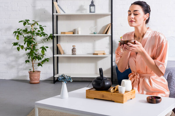 woman in silk bathrobe with wooden bowl in hands having tea ceremony in morning at home