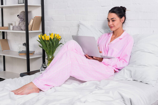 smiling woman in pajamas using laptop in bed at home