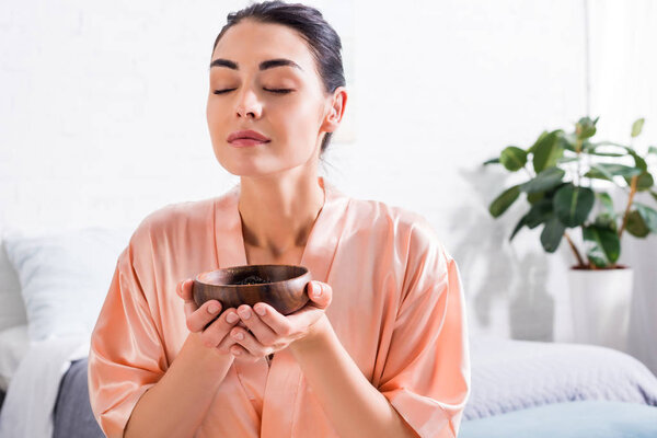 woman in silk bathrobe with wooden bowl in hands having tea ceremony in morning at home