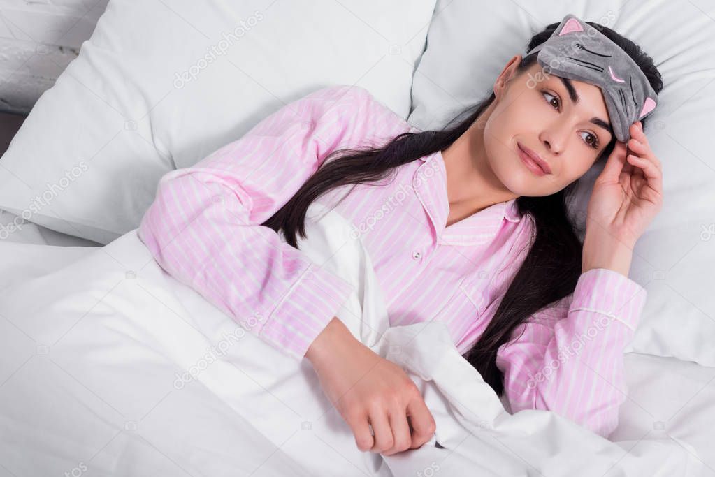 portrait of pensive woman in pink pajamas and sleeping mask lying in bed at home