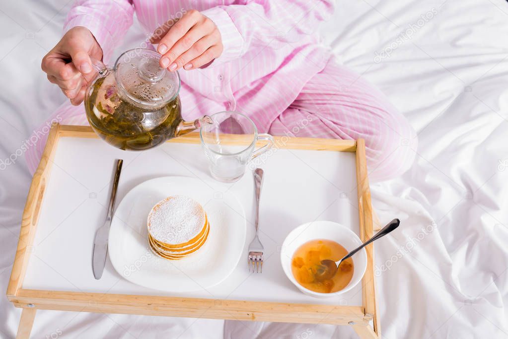 partial view of woman in pink pajamas pouring green tea into cup while having breakfast in bed at home