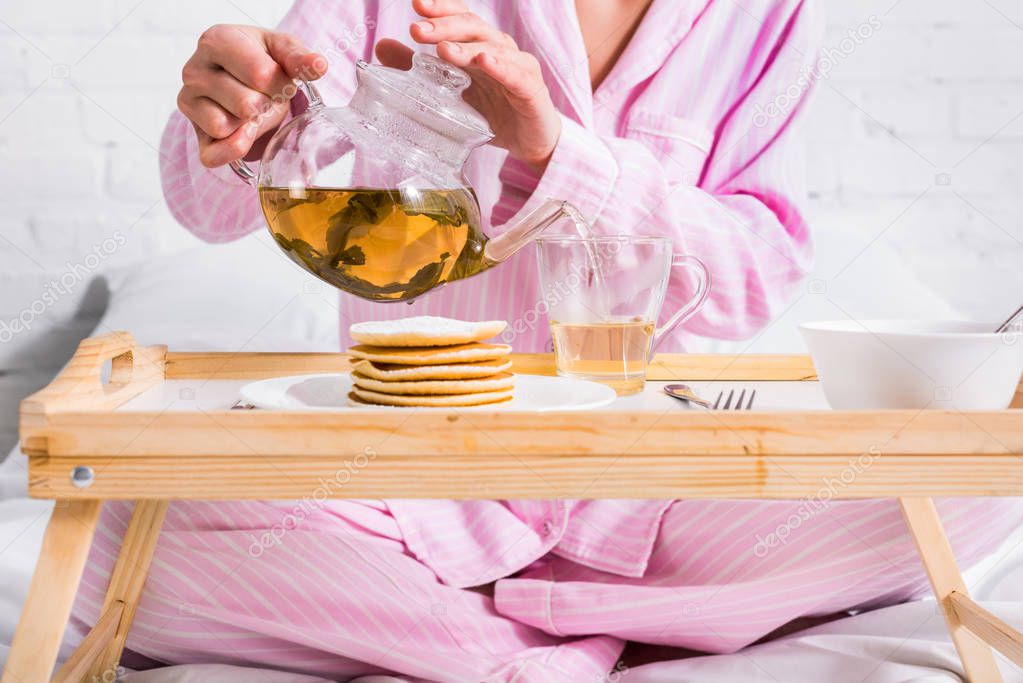 partial view of woman in pink pajamas pouring green tea into cup while having breakfast in bed at home