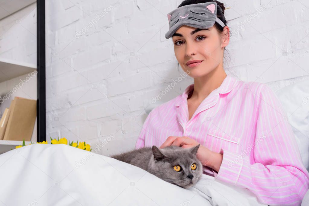 portrait of woman in pajamas and sleeping mask with britain shorthair cat resting in bed at home