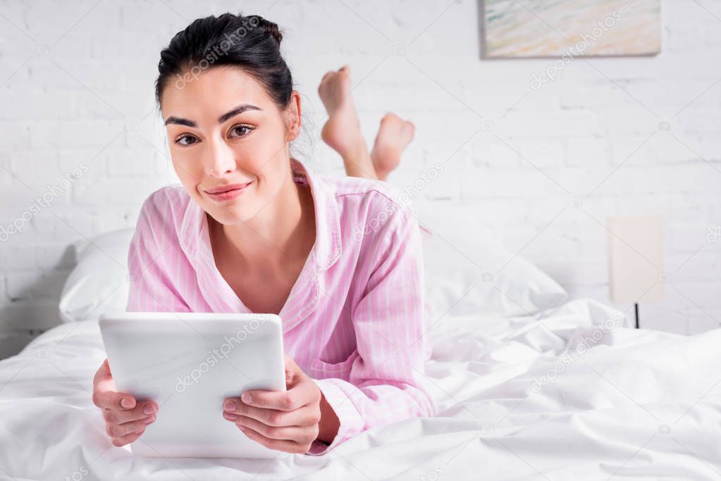 portrait of smiling woman in pajamas with tablet while lying on bed at home