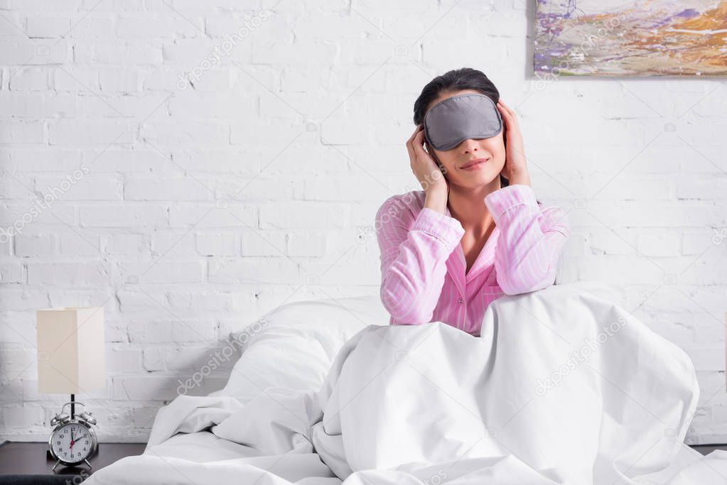 woman in pink pajamas and sleeping mask sitting on bed at home