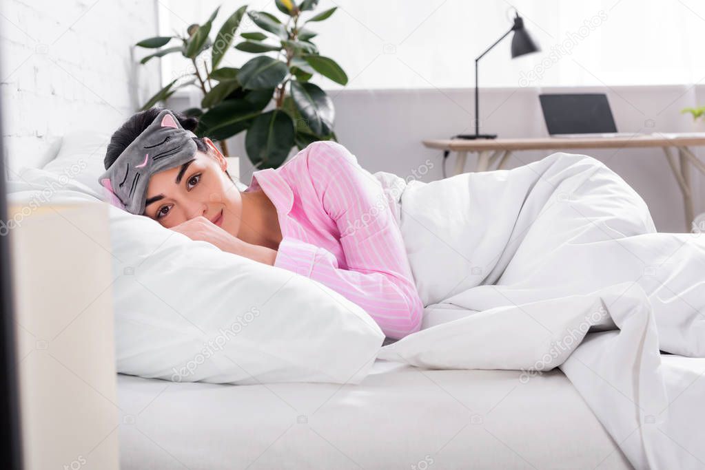 portrait of woman in pink pajamas and sleeping mask lying in bed at home