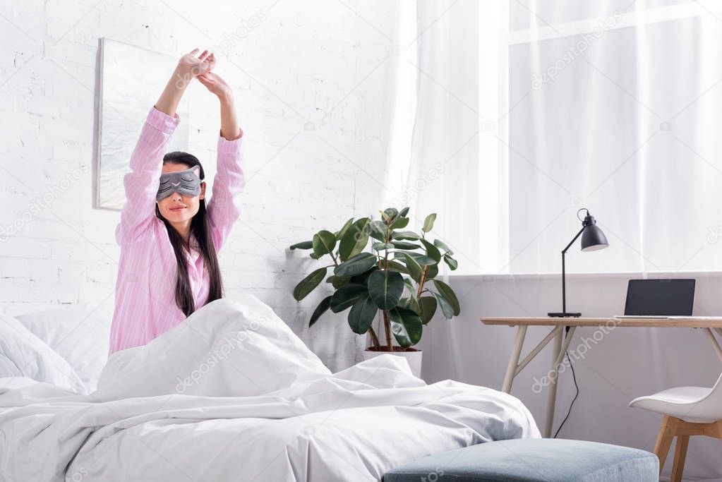 portrait of woman in pink pajamas and sleeping mask stretching in bed in morning
