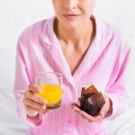 Cropped shot of woman in pink pajamas with glass of fresh juice and chocolate muffin in hands on bed at home