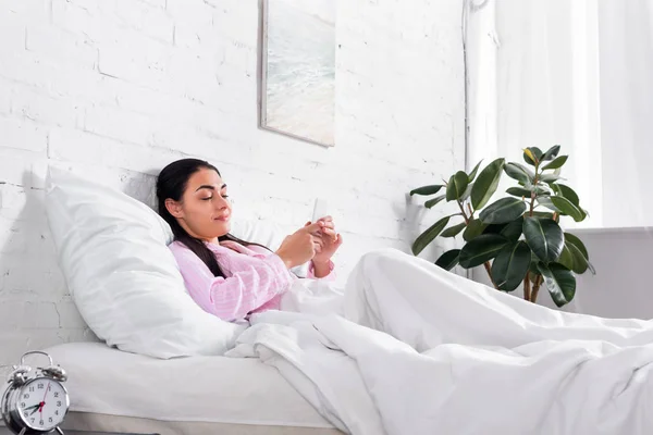 穿着睡衣的微笑的女人在家里睡觉时使用智能手机 — 图库照片