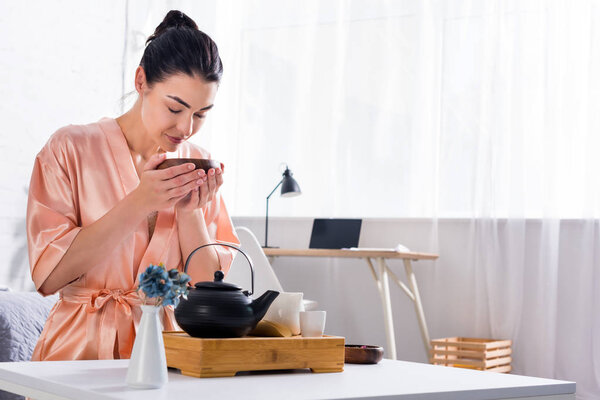 woman in silk bathrobe with wooden bowl in hands having tea ceremony in morning at home