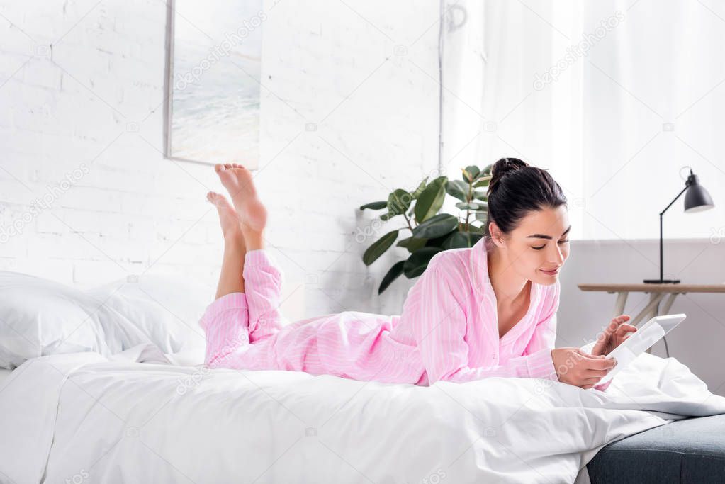 side view of smiling woman in pajamas using tablet while lying on bed at home