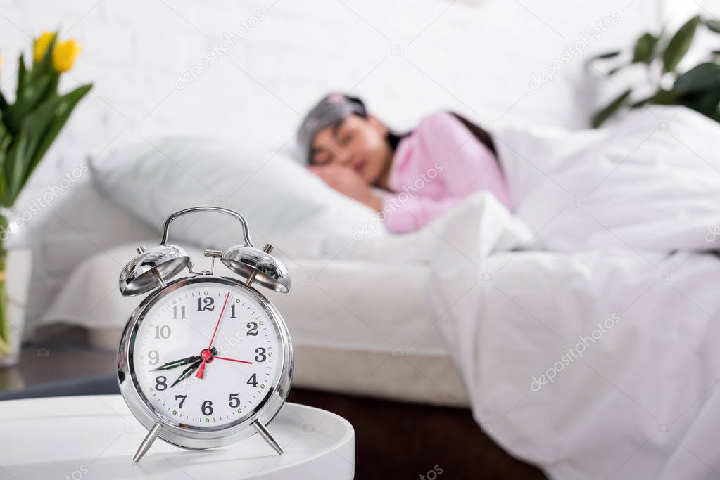 selective focus of clock and woman in pink pajamas and sleeping mask sleeping in bed at home