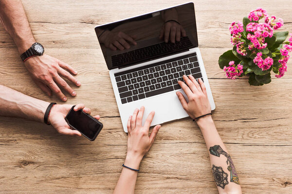 cropped shot of man with smartphone in hand and woman at tabletop with laptop and kalanchoe flower