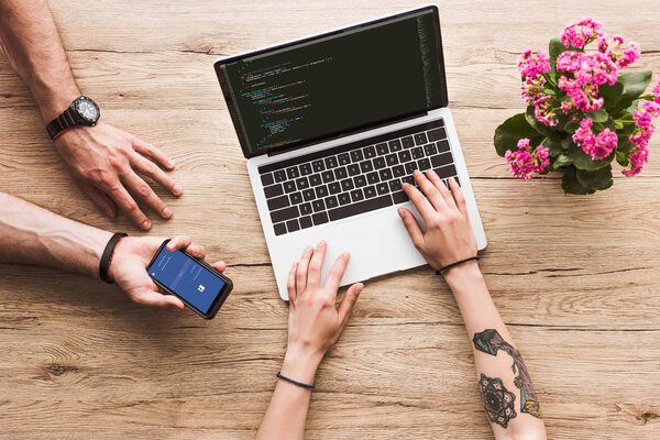 обрезанный снимок мужчины со смартфоном с логотипом facebook в руке и женщиной за столом с ноутбуком и цветком каланхое
