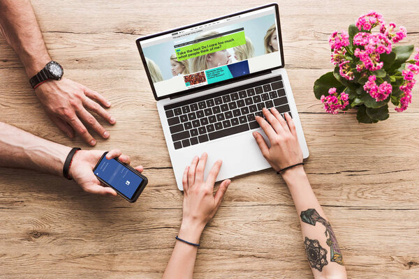 cropped shot of man with smartphone with facebook logo in hand and woman at tabletop with laptop with bbc website and kalanchoe flower