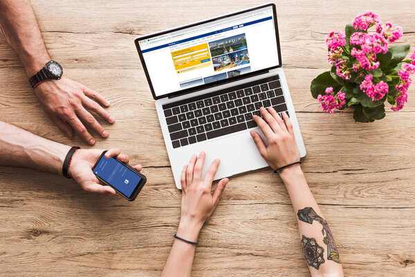 cropped shot of man with smartphone with facebook logo in hand and woman at tabletop with laptop with bookingcom website and kalanchoe flower