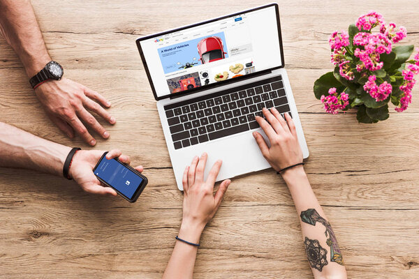 cropped shot of man with smartphone with facebook logo in hand and woman at tabletop with laptop with ebay website and kalanchoe flower