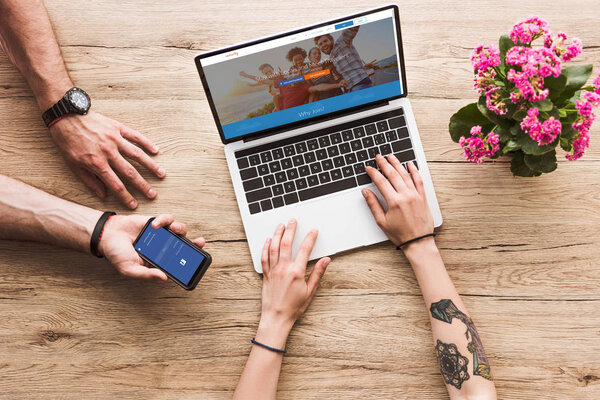 cropped shot of man with smartphone with facebook logo in hand and woman at tabletop with laptop with couchsurfing website and kalanchoe flower
