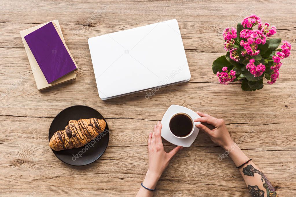 cropped shot of woman with cup of coffee at tabletop with laptop, books and croissant on plate