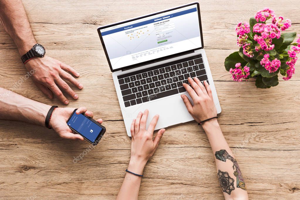 Cropped shot of man with smartphone with facebook logo in hand and woman at tabletop with laptop facebook website and kalanchoe flower
