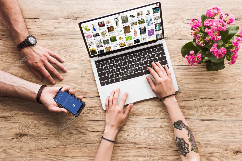 Cropped shot of man with smartphone with facebook logo in hand and woman at tabletop with laptop with pinterest website and kalanchoe flower