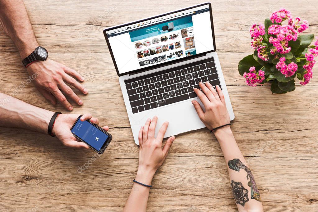 Cropped shot of man with smartphone with facebook logo in hand and woman at tabletop with laptop with amazon website and kalanchoe flower