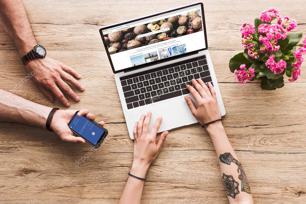 Cropped shot of man with smartphone with facebook logo in hand and woman at tabletop with laptop with depositphotos website and kalanchoe flower
