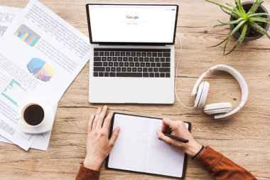 bir google logosu, kağıtları, fincan kahve ve kulaklık ile dizüstü bilgisayar ile işyerinde Not defterinizdeki notlar yapma adam kısmi görünümü