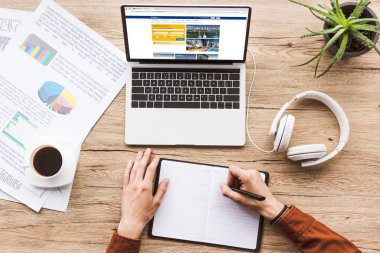 kısmi bookingcom logo, kağıtları, fincan kahve ve kulaklık ile dizüstü bilgisayar ile işyerinde Not defterinizdeki notlar yapma adam görünümünü