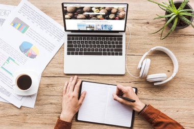 laptop depositphotos Web sitesi, kulaklık, ders kitabı, kalem, infographics, kahve fincanı ve masada saksılı bitki ile çalışan adamın kısmi görünümü