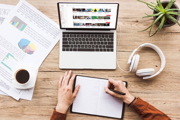 partial view of man making notes in notebook at workplace with laptop with youtube website, papers, cup of coffee and headphones