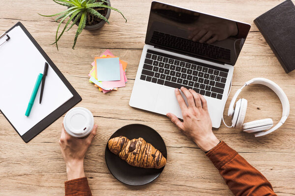 cropped image of man working at table with laptop, headphones, textbook, clipboard, post it, potted plant, coffee cup and croissant 