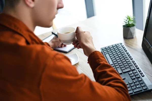 コーヒーを飲むと コンピューターとコンピューターのキーボードを持つテーブルで教科書を書く男のショットをトリミング — ストック写真