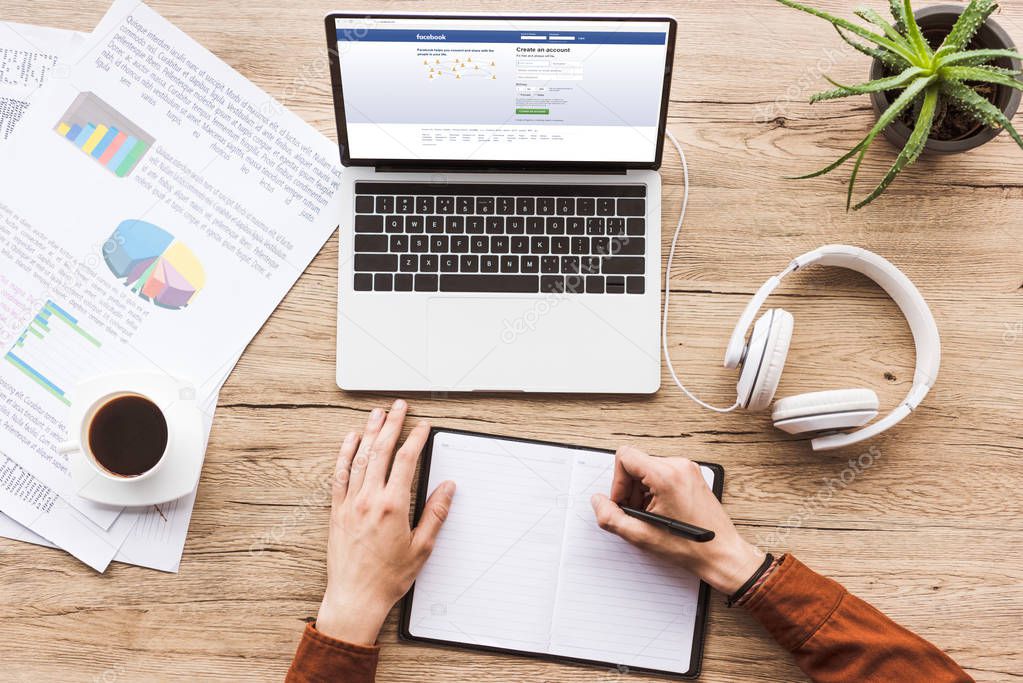 Partial view of man making notes in notebook at workplace with laptop with facebook logo, papers, cup of coffee and headphones